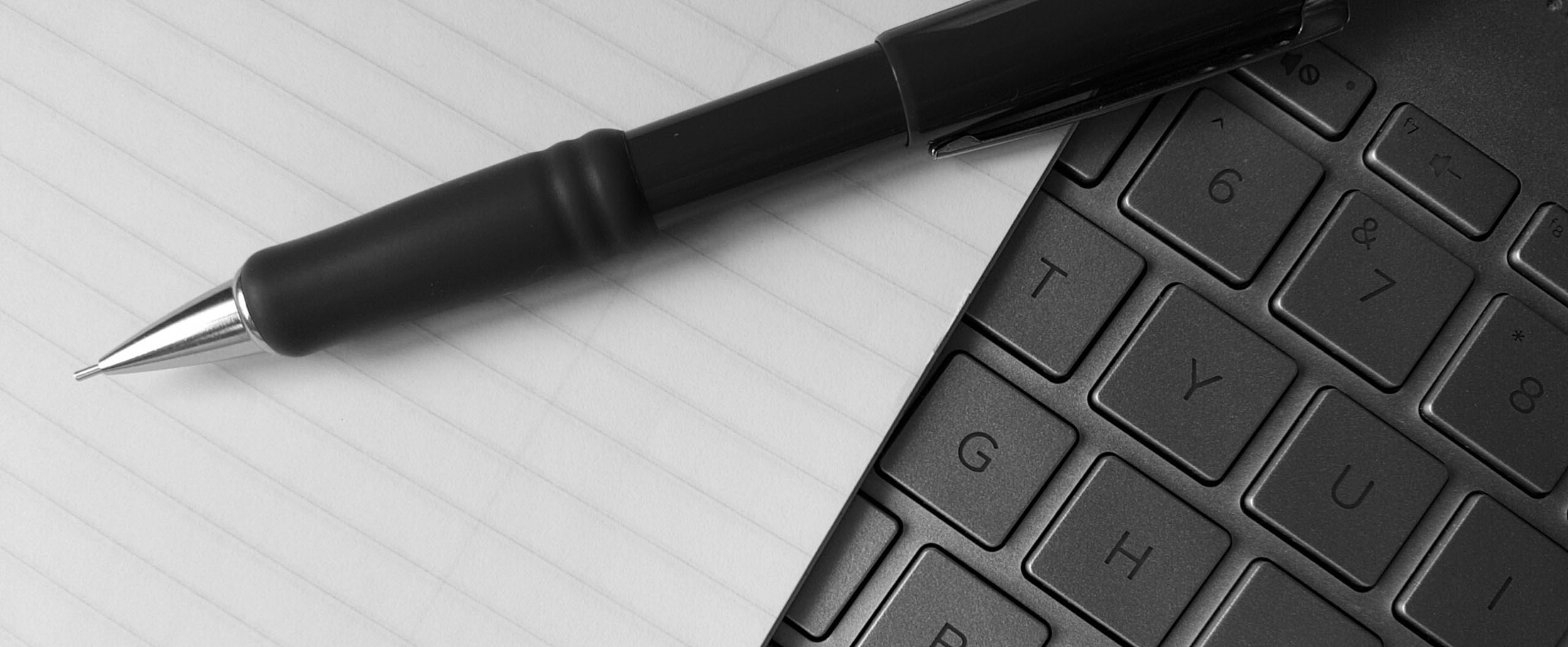 black and white close-up of a mechanical pencil resting on lined paper atop a computer keyboard